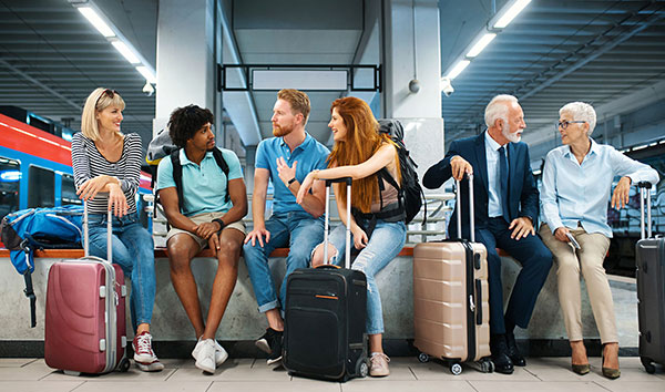 Sechs Personen, jung und alt, sitzen an einem Bahnhof mit ihren Koffern und unterhalten sich